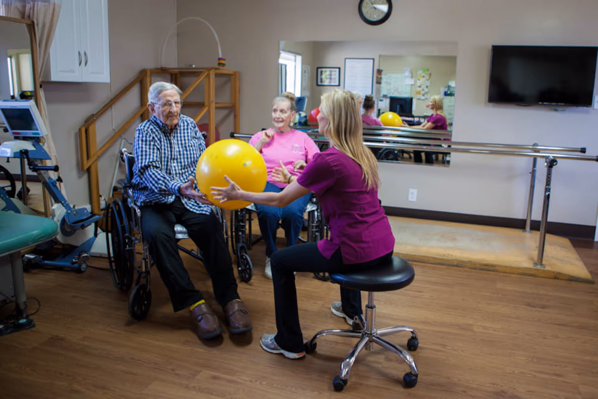 A physical therapy session in a rehabilitation room where a female therapist in a purple uniform is assisting two elderly individuals in wheelchairs with an exercise using a large yellow therapy ball. The room has wooden floors, a wall-mounted TV, a large mirror, and parallel bars for walking exercises.