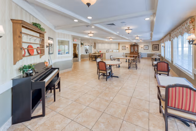 A spacious, well-lit common area with tiled floors and multiple tables and chairs arranged for seating. On the left side, there is a black upright piano with a bench, and above it, a wooden wall shelf with decorative plates and greenery. The room has large windows on the right side with floral valances, allowing natural light to fill the space. The ceiling features recessed lighting and hanging light fixtures. The far end of the room has a counter area and a wooden cabinet with framed pictures on the walls.