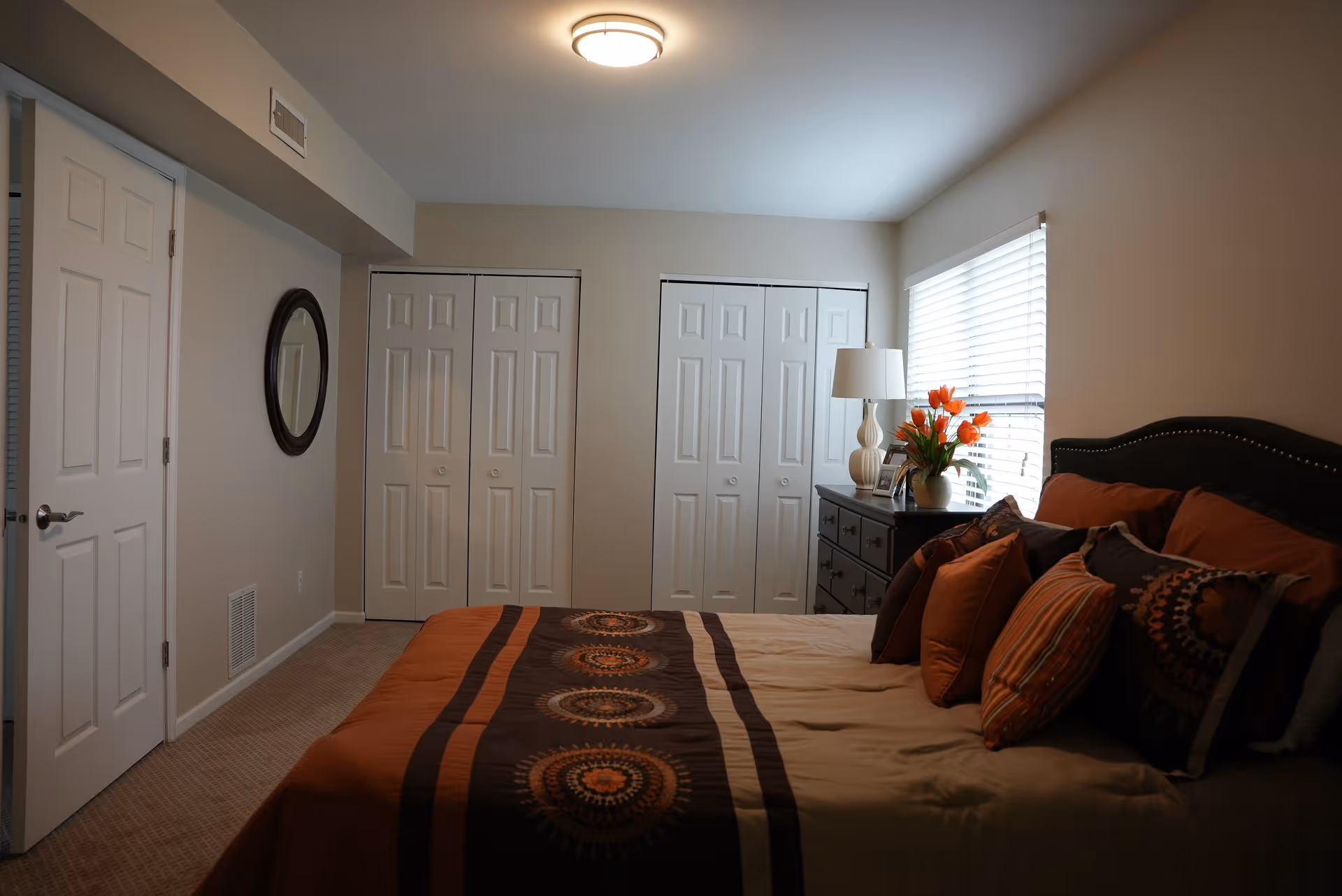 Well-lit bedroom with a bed dressed in orange and brown linens, a dresser with a lamp and flowers, and double closet doors.