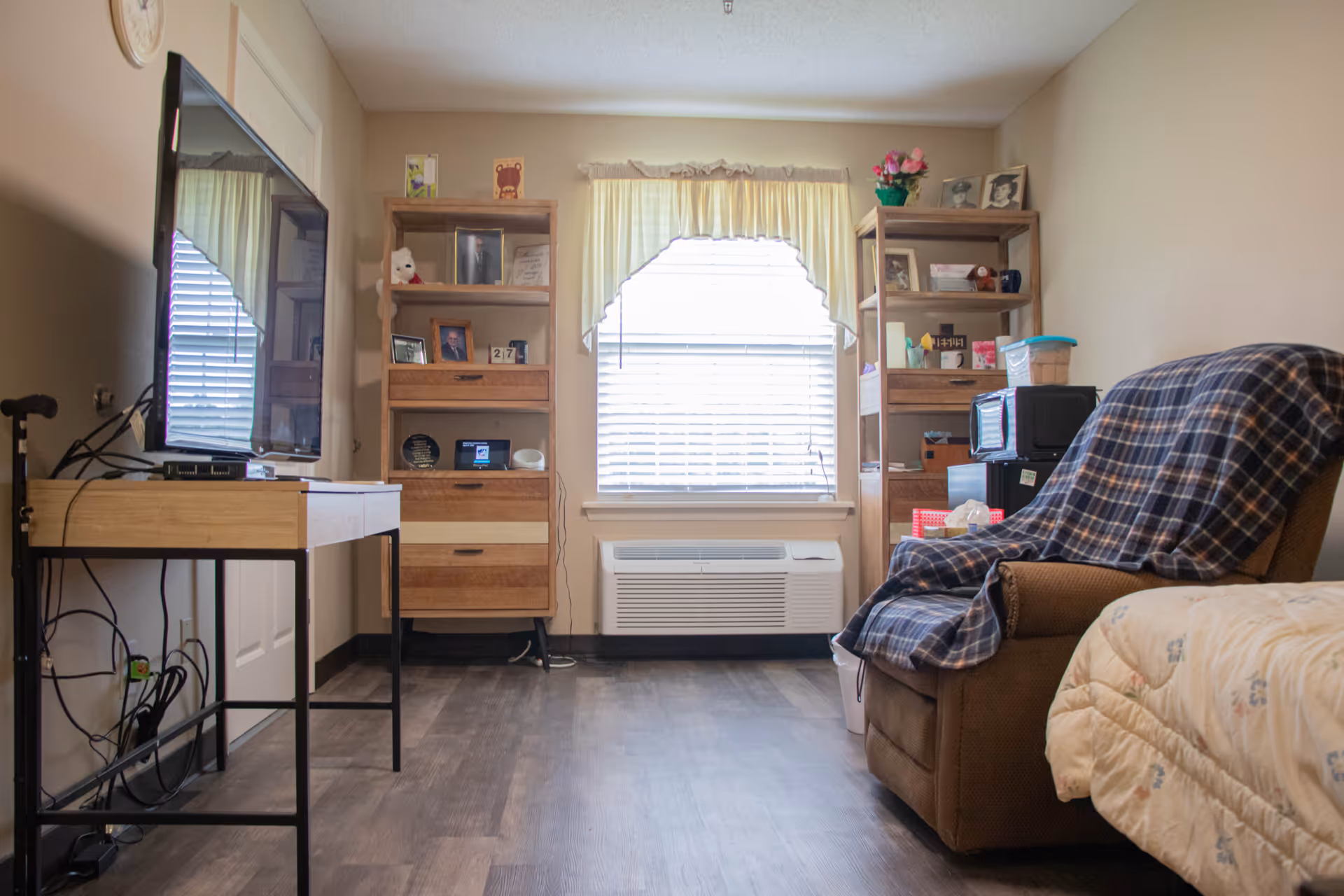 A cozy room with a brown recliner covered by a plaid blanket, a bed with a floral comforter, two wooden shelving units filled with framed photos and various items, a window with blinds and a valance, and a flat-screen TV mounted on a light wood table with black metal legs. The floor is dark wood, and there is an air conditioning unit below the window.