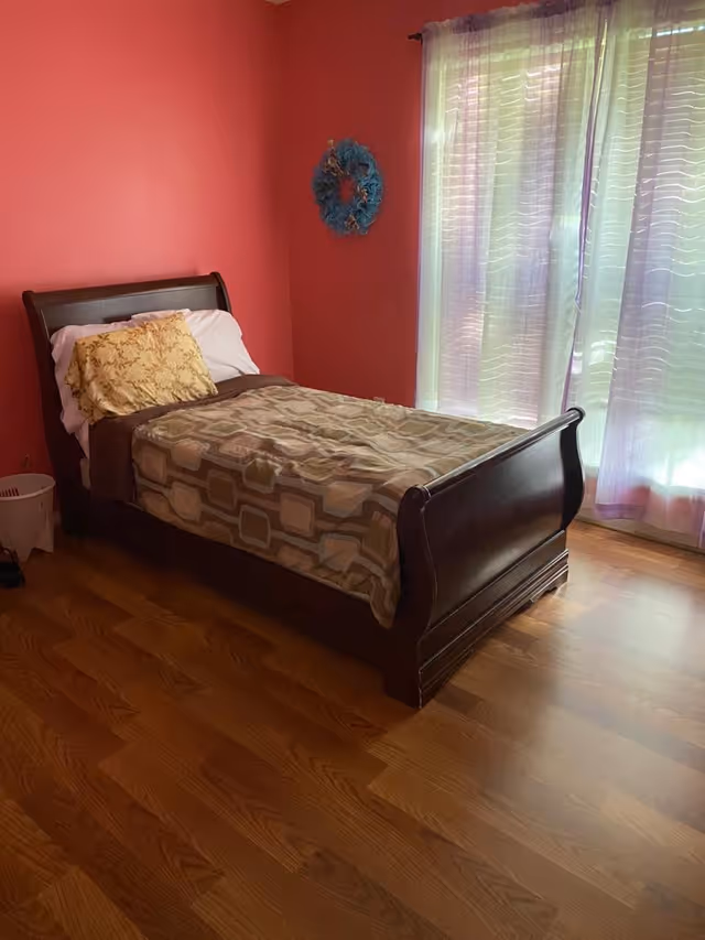 Single wooden sleigh bed with pillows and patterned blanket in a pink-walled bedroom next to a sheer-curtained window on hardwood floors.