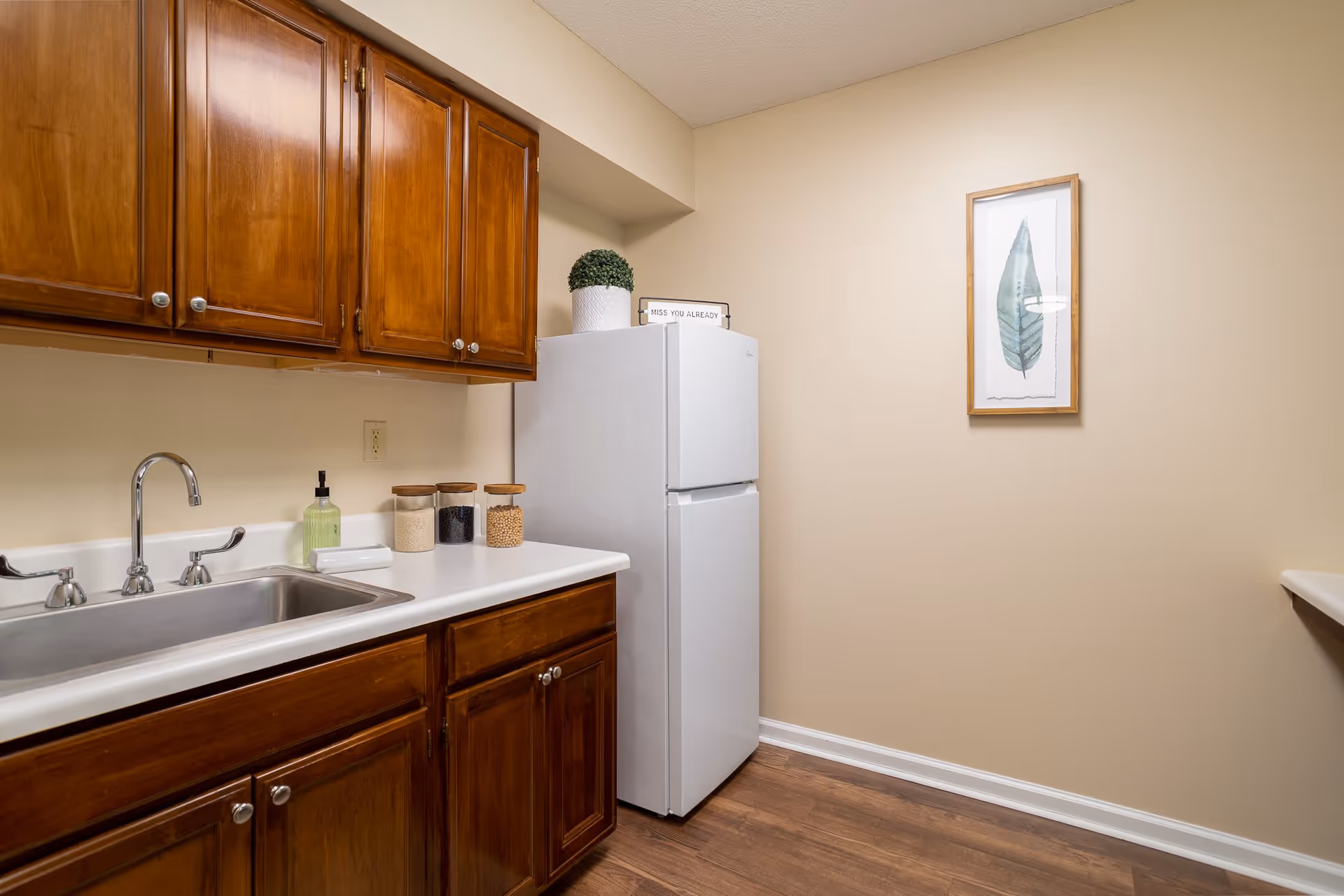 A small kitchen area with wooden cabinets, a white countertop, a stainless steel sink with a faucet, and a white refrigerator. On the countertop are three glass jars with wooden lids and a soap dispenser. A small potted plant and a sign that reads 'MISS YOU ALREADY' are placed on top of the refrigerator. A framed picture of a green leaf hangs on the beige wall.