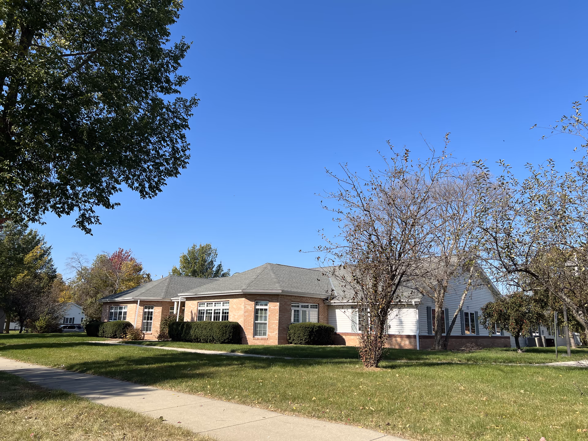 Exterior view of a single-story senior living facility building with brick and siding walls, surrounded by green grass, trees, and a sidewalk under a clear blue sky.
