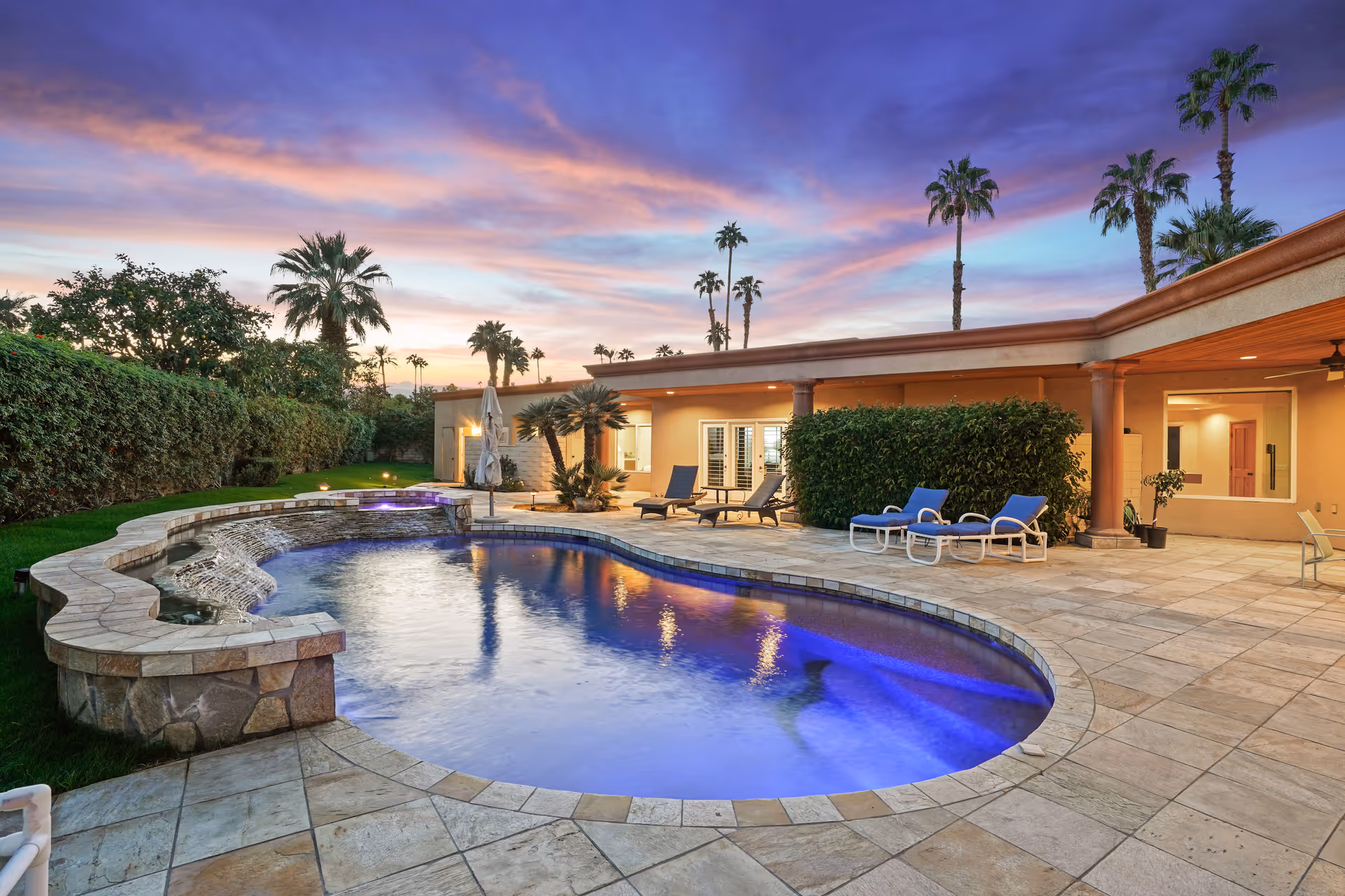 A luxurious outdoor pool area at sunset with a uniquely shaped swimming pool illuminated with blue lights. The pool is surrounded by a tiled patio with lounge chairs and greenery, including palm trees and bushes. The background shows a single-story building with large windows and a covered patio area.