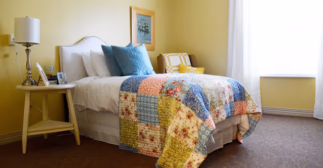 A cozy bedroom with a bed covered in a colorful patchwork quilt, white pillows, and blue accent pillows. There is a small round bedside table with a lamp, picture frames, and a small clock. A framed picture hangs on the yellow wall above the bed, and a window with white curtains allows natural light into the room. A yellow and white checkered chair is placed near the window.
