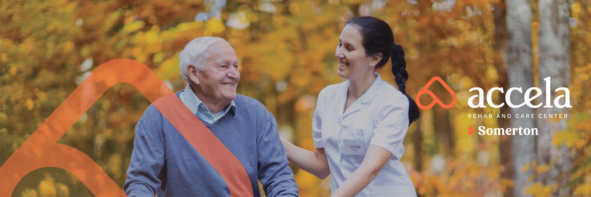 An elderly man in a blue sweater is smiling while being assisted by a smiling female caregiver in a white uniform outdoors with autumn foliage in the background.