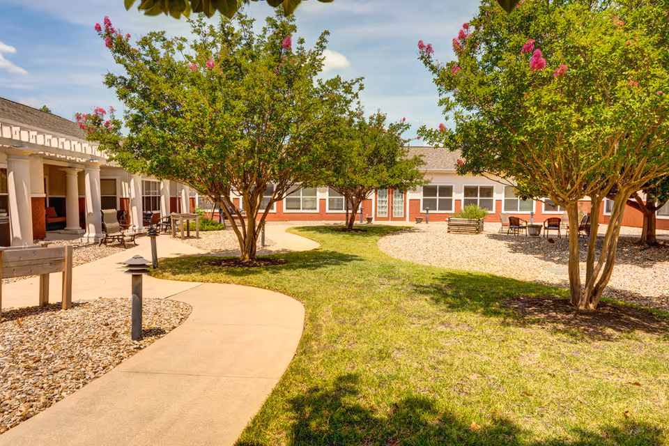 Outdoor courtyard area at Parmer Woods at North Austin featuring a curved concrete walkway, green grass, trees with pink flowers, and seating areas with chairs and tables. The building with large windows and a covered porch with columns surrounds the courtyard.