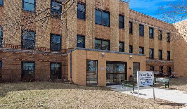 Exterior view of a three-story brick building with multiple windows and a main entrance with glass doors. There are two green benches near the entrance and a sign that reads 'Baker Park Community Housing, Senior Living & Parking, Sanford Health'. Leafless tree branches are visible in the foreground under a clear blue sky.