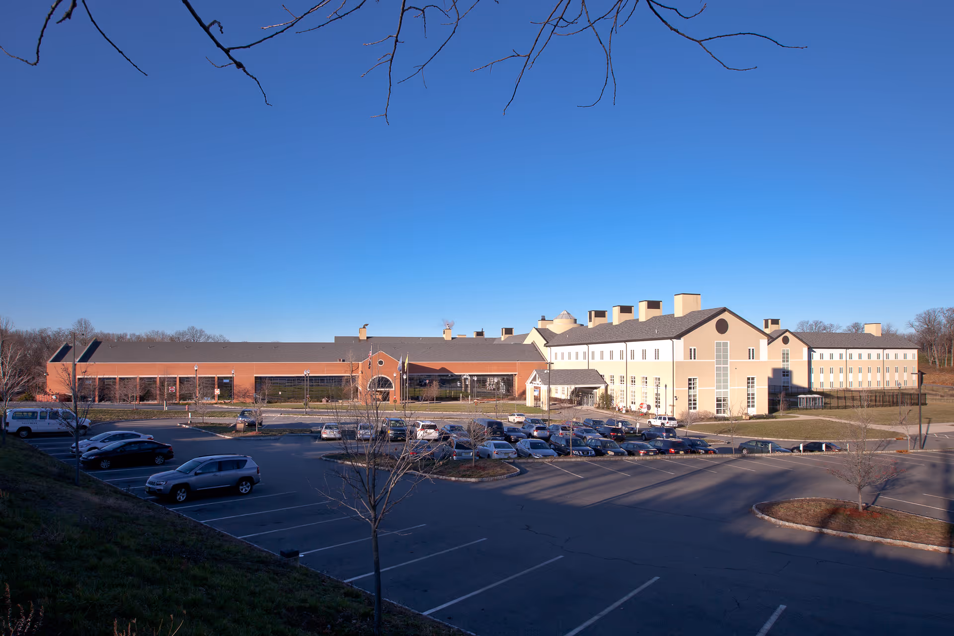 Exterior view of Preakness Healthcare Center, a large multi-story building with a parking lot in front. Several cars are parked, and the sky is clear and blue.