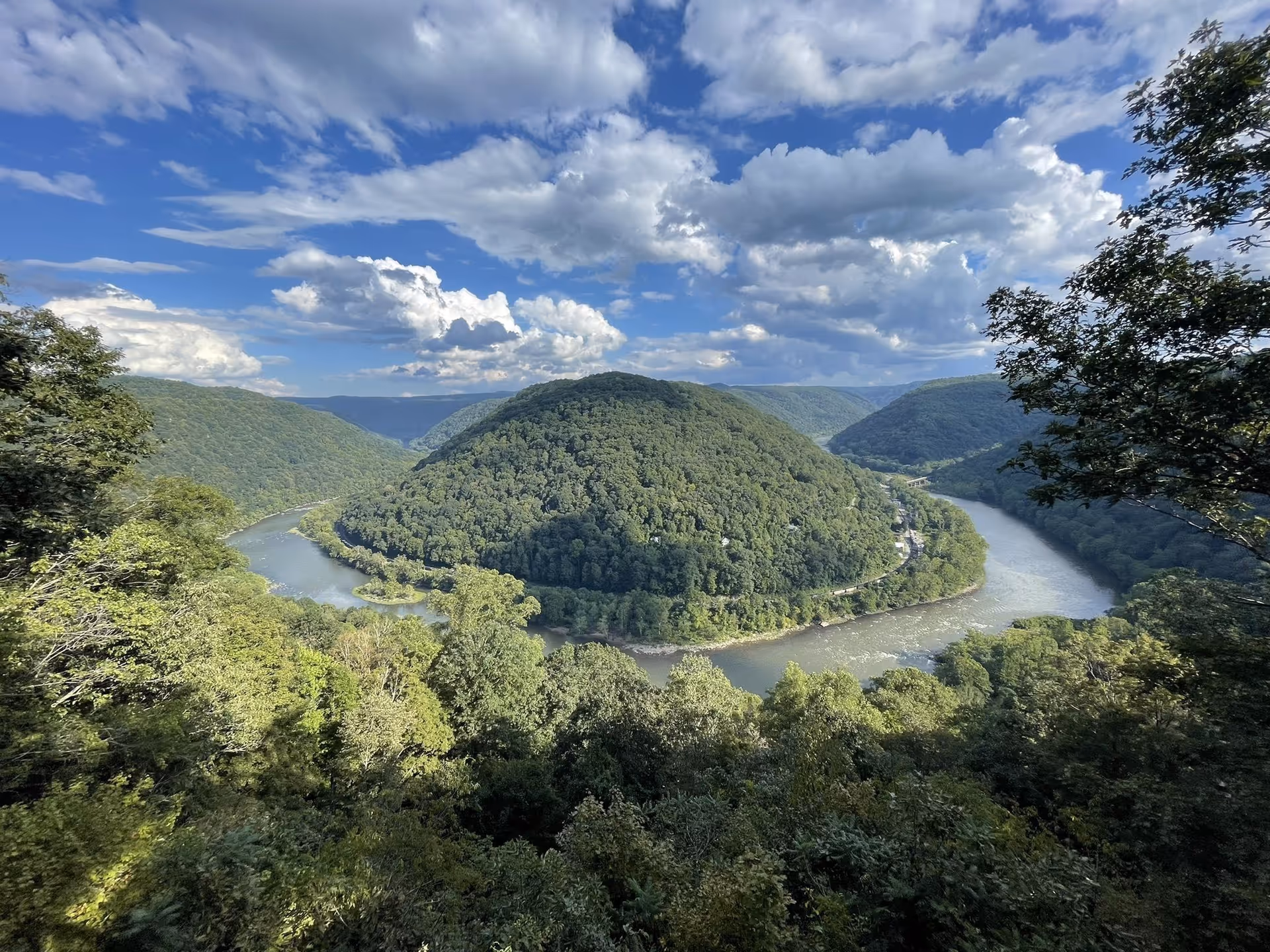 A scenic view of a river winding around a densely forested hill under a partly cloudy blue sky, with lush green trees in the foreground.