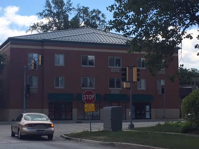 Three-story brick apartment building with a metal roof at a street intersection, traffic lights, a stop sign, and a car in front.