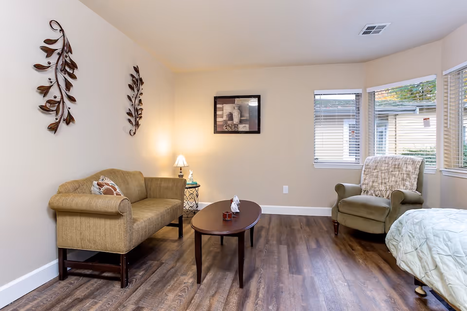 Bright sitting area with a sofa, armchair, coffee table, side table with lamp, wall art and windows in a senior living room.