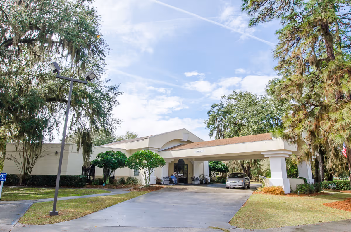 Exterior view of Surrey Place Nursing Center showing the entrance with a covered driveway. There are trees and shrubs around the building, a car parked under the covered entrance, and a clear blue sky with some clouds.
