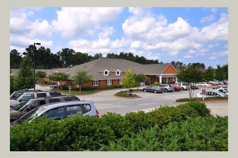 Exterior view of a single-story brick senior living building with a parking lot, cars, and landscaped shrubs under a partly cloudy sky.