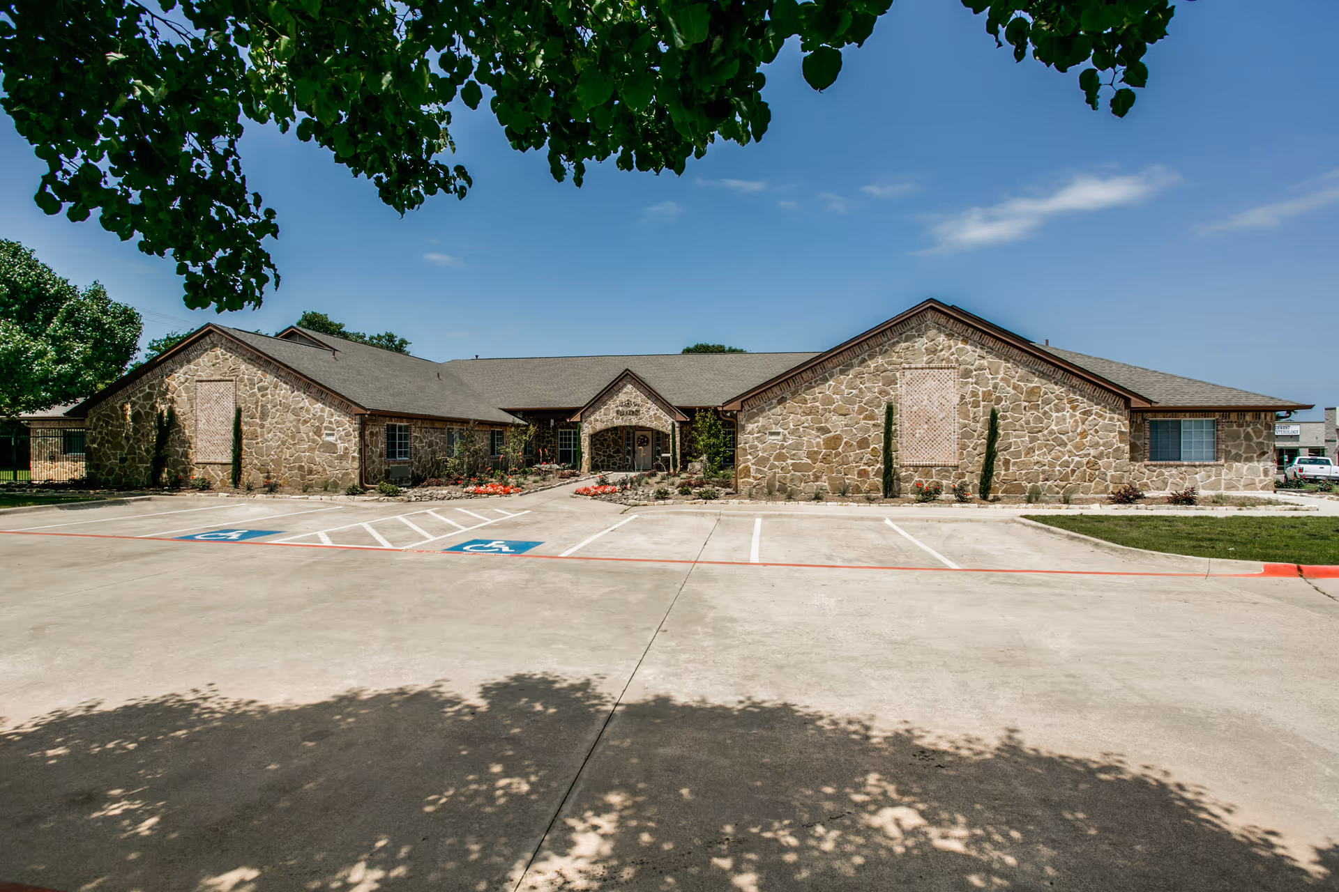 Front exterior view of a single-story stone building with a pitched roof, surrounded by a parking lot with two handicap parking spaces marked. There are small landscaped areas with shrubs and flowers near the entrance, and trees partially frame the top of the image under a clear blue sky.