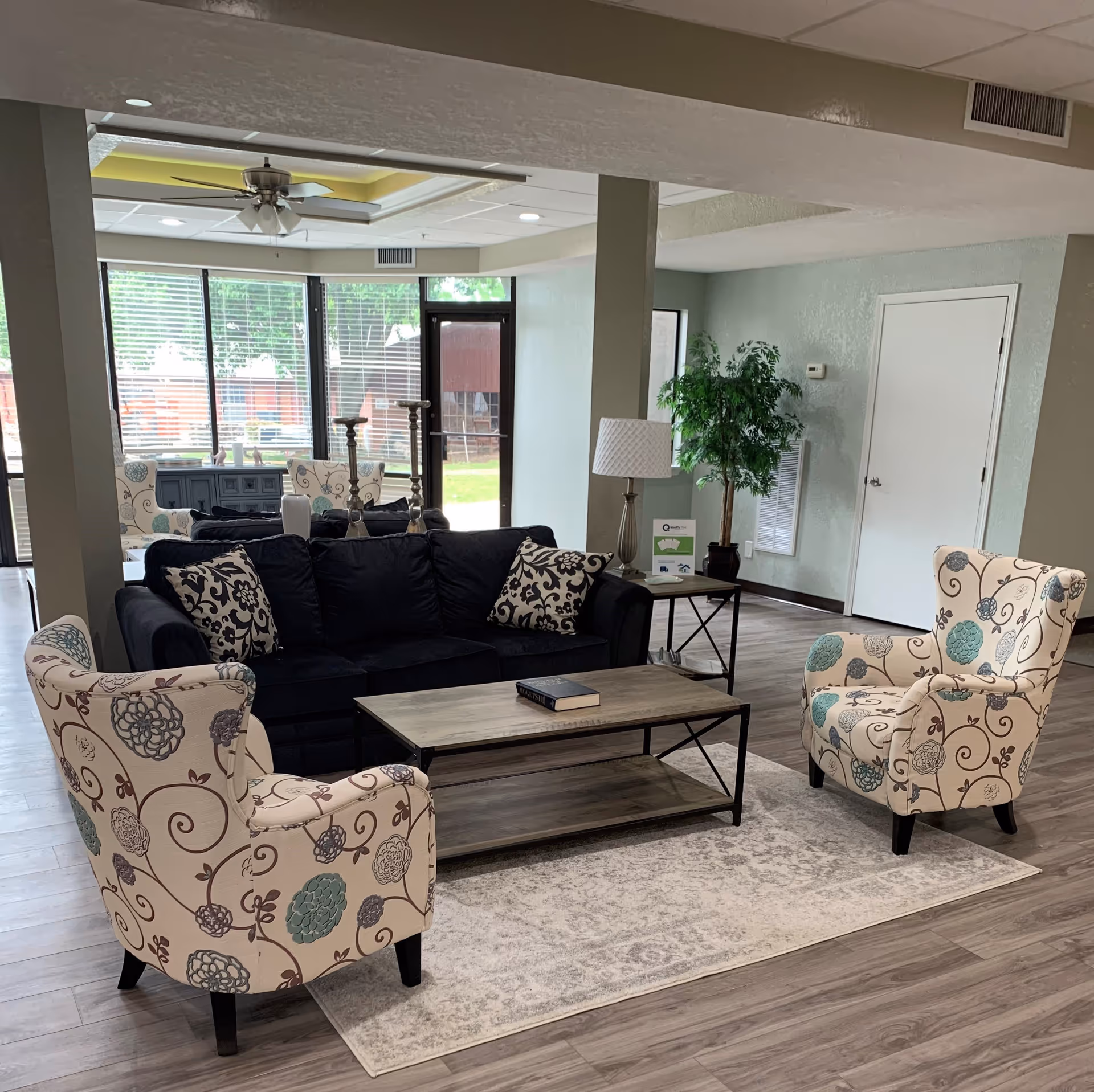 A cozy living room area with a black sofa adorned with patterned pillows, two floral-patterned armchairs, a wooden coffee table with a book on it, a side table with a lamp, and a potted plant. Large windows and a glass door provide natural light and a view of the outdoor greenery.