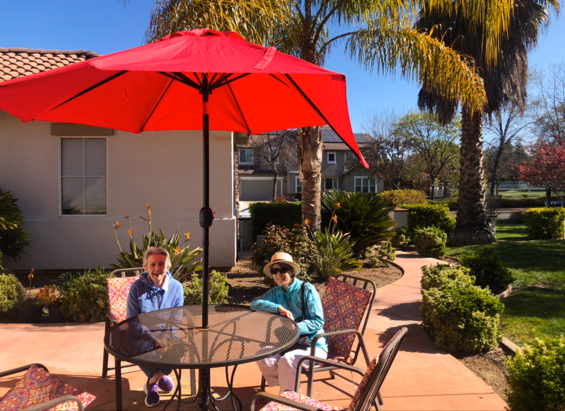 Two elderly women sitting at a round outdoor metal table with a large red umbrella providing shade. They are in a garden area with palm trees, bushes, and a paved walkway. The weather is sunny with a clear blue sky.