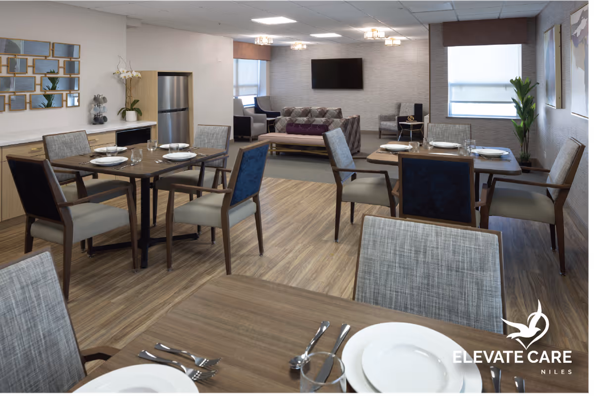 A dining area in a senior living facility with several wooden tables set with white plates, glasses, and silverware. The room has wooden flooring and modern chairs with fabric upholstery. In the background, there is a living room area with a sofa, armchairs, a wall-mounted TV, and large windows with blinds. A small kitchenette with a refrigerator and cabinets is visible on the left side. The room is well-lit with ceiling lights and decorated with plants and wall art.