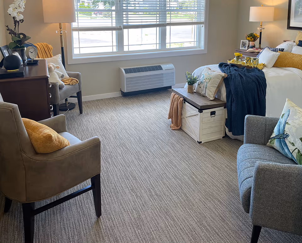A cozy bedroom with a large window letting in natural light. The room features a bed with white and yellow bedding, a wooden chest at the foot of the bed with a blue blanket and decorative pillows, two armchairs with cushions, a wooden dresser with a lamp and clock, and a floor lamp beside a gray armchair. The carpet is light-colored and the walls are painted beige.