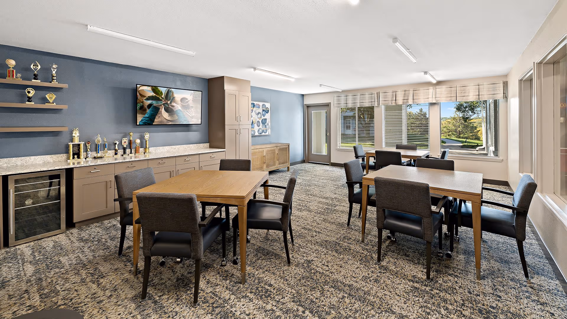 A bright and spacious common area with multiple wooden tables and cushioned chairs arranged for seating. The room features a blue accent wall with shelves displaying trophies and a framed picture. Large windows with blinds allow natural light to fill the space, offering a view of greenery outside. The carpet has a patterned design, and there are cabinets and a small refrigerator along one wall.