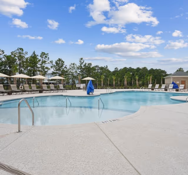 Outdoor swimming pool area with clear blue water, surrounded by lounge chairs and umbrellas. Trees and greenery are visible in the background under a partly cloudy sky.