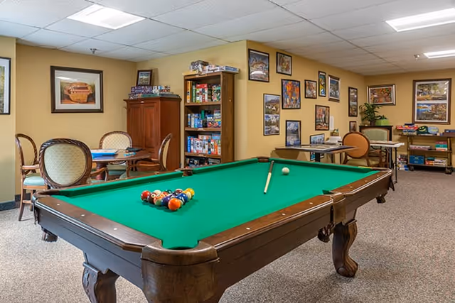 A recreational room with a green felt pool table in the foreground, set up for a game with balls racked and a cue stick resting on the table. In the background, there are several tables and chairs, shelves filled with board games, framed pictures on the walls, and a carpeted floor.