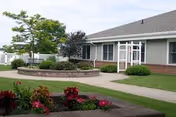 Single-story senior living building front with a paved walkway, landscaped flower beds, and a small courtyard trellis.