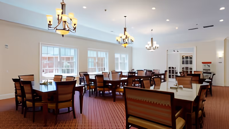 Well-lit dining room with multiple wooden tables and chairs beneath chandeliers and large windows.
