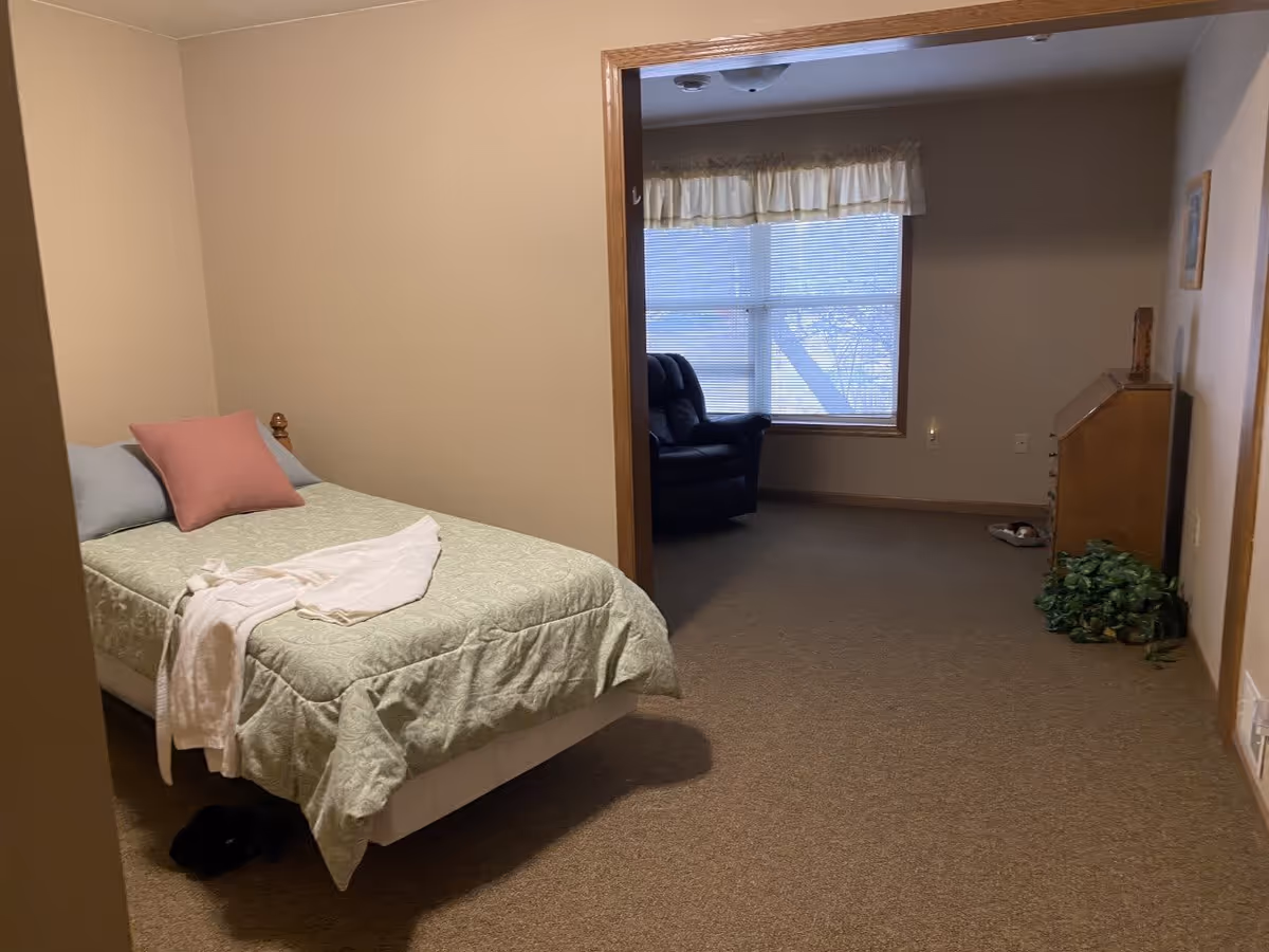 A small bedroom with a single bed covered in a light green quilt and two pillows, one pink and one blue. A white towel is draped over the bed. The room has beige walls and carpet. Through an open doorway, a living area is visible with a black recliner chair, a window with blinds and a valance, a wooden dresser, a small plant on the floor, and a framed picture on the wall.
