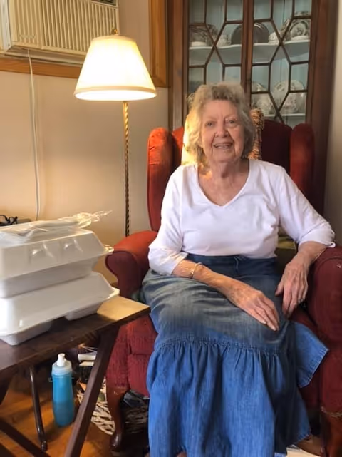 An elderly woman with gray hair wearing a white long-sleeve shirt and a blue skirt is sitting in a red armchair. Next to her is a wooden side table with two stacked white takeout containers and a blue water bottle on the floor. A floor lamp with a beige shade is behind the table, and a glass-front cabinet with decorative plates is in the background.