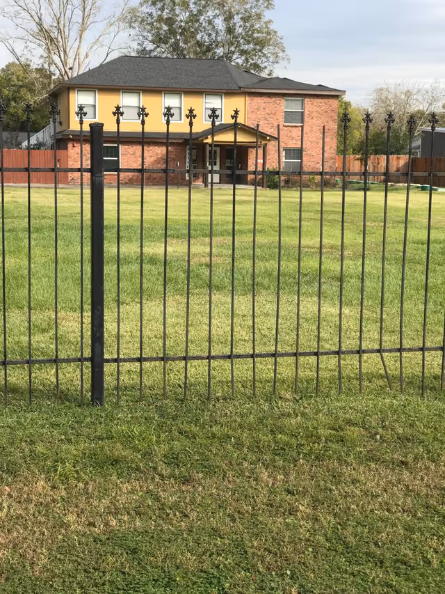View of a two-story brick and yellow house with a black roof, seen through a black metal fence. The house is surrounded by a large green lawn and some trees in the background.