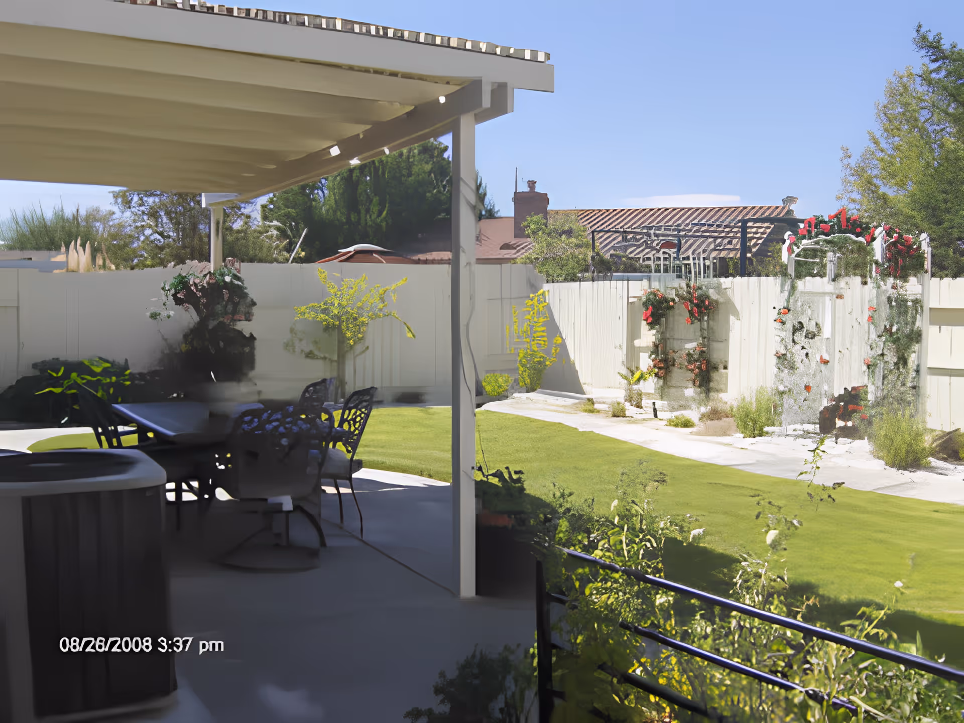 A covered patio with an outdoor table and chairs opening onto a grassy backyard with a white fence and flowering trellis.