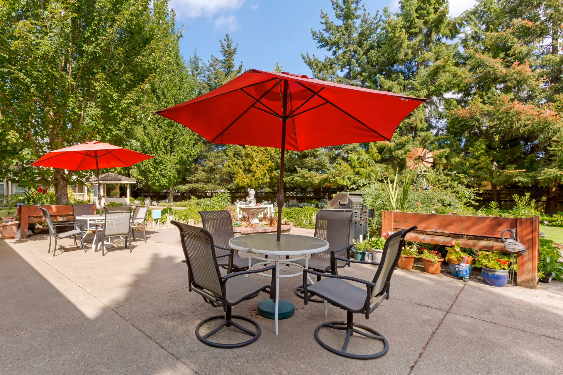 Outdoor patio area with round tables and chairs under large red umbrellas, surrounded by greenery including trees and potted plants, with a clear blue sky overhead.