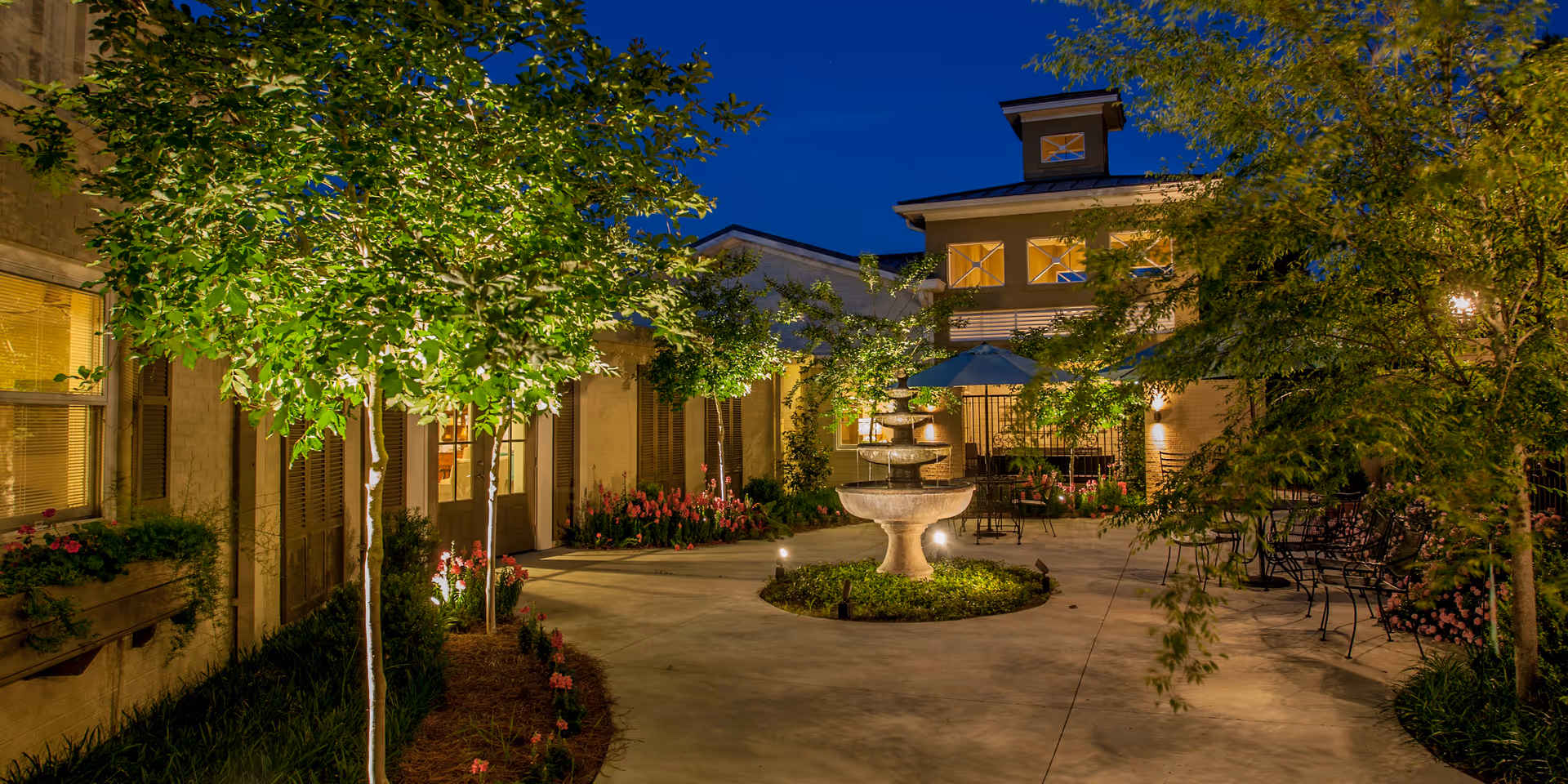 A well-lit outdoor courtyard at night featuring a central multi-tiered stone fountain surrounded by greenery and flowering plants. The courtyard is enclosed by a building with windows and doors, and there are several metal chairs and tables with umbrellas arranged around the space. Trees and shrubs add to the serene atmosphere.