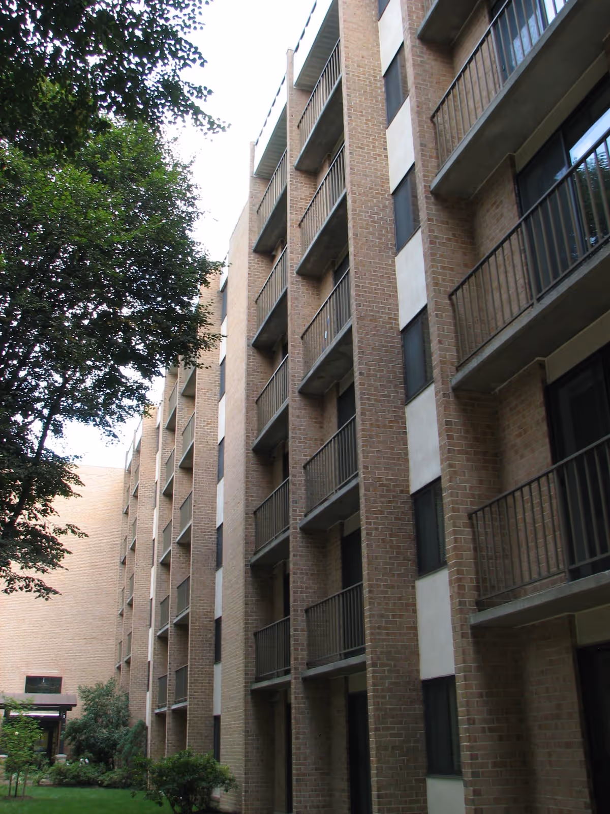 Multi-story brick apartment building with stacked balconies overlooking a grassy courtyard and trees.