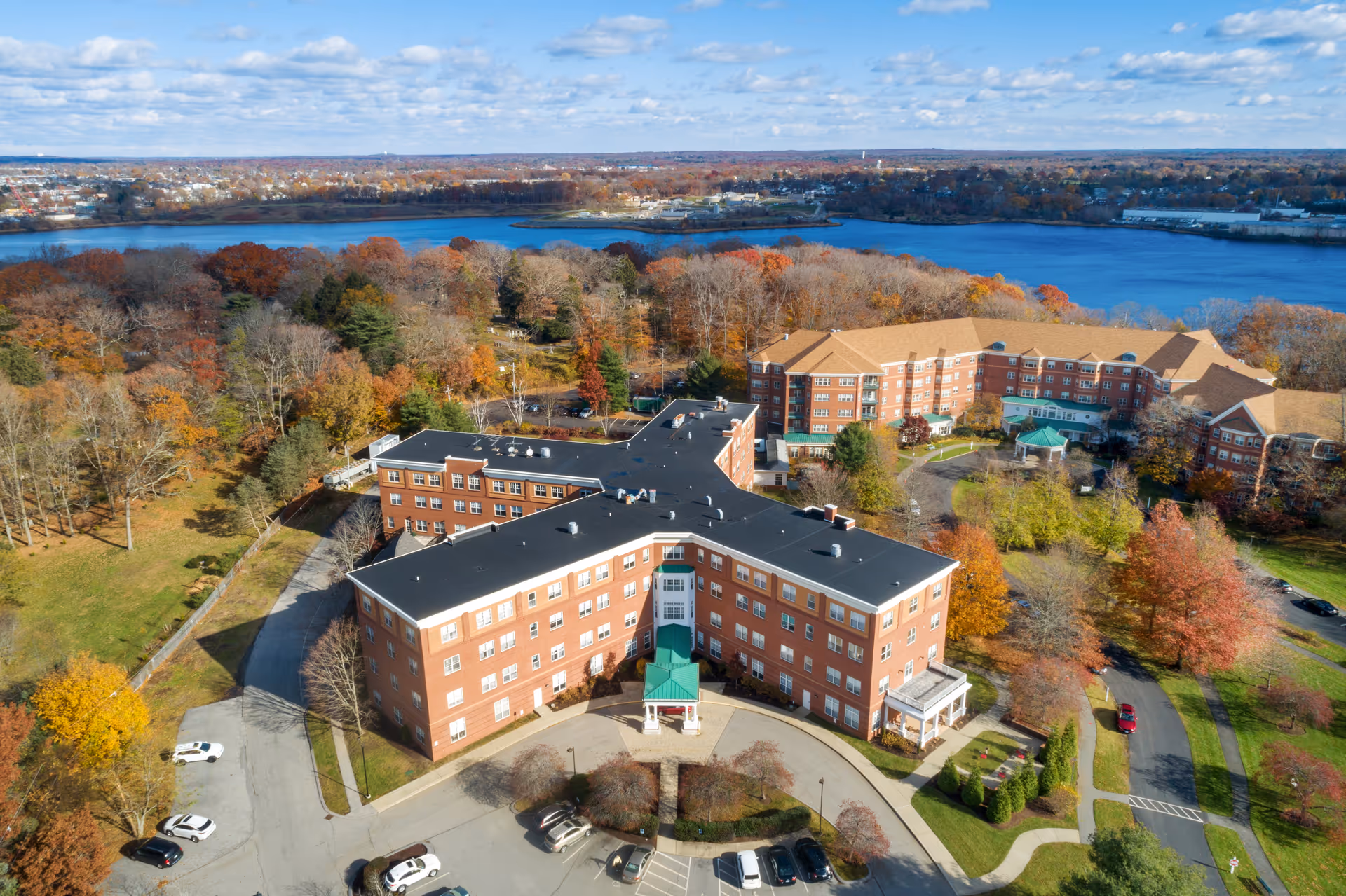 Aerial view of Wingate Residences on Blackstone Boulevard, showing a large multi-story brick building complex surrounded by trees with autumn foliage. The complex is near a large body of water under a partly cloudy sky. Several cars are parked in the parking lot in front of the building.