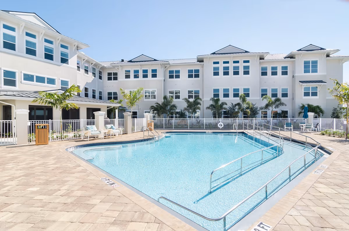 Outdoor swimming pool area at American House Ft. Myers with clear blue water, surrounded by a tiled deck, white safety railings, palm trees, and a multi-story white building in the background under a clear blue sky.