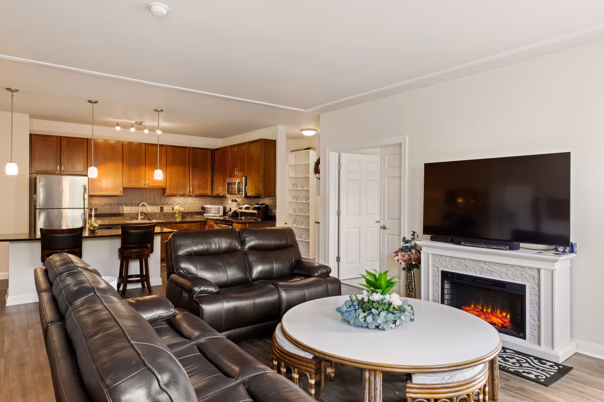 A modern living room with two black leather sofas facing a white round coffee table decorated with a green and white floral arrangement. A flat-screen TV is mounted above a white electric fireplace on the right wall. In the background, there is an open kitchen with wooden cabinets, stainless steel appliances, and three pendant lights hanging over a breakfast bar with two dark wooden chairs. The room has light-colored walls and wood flooring.