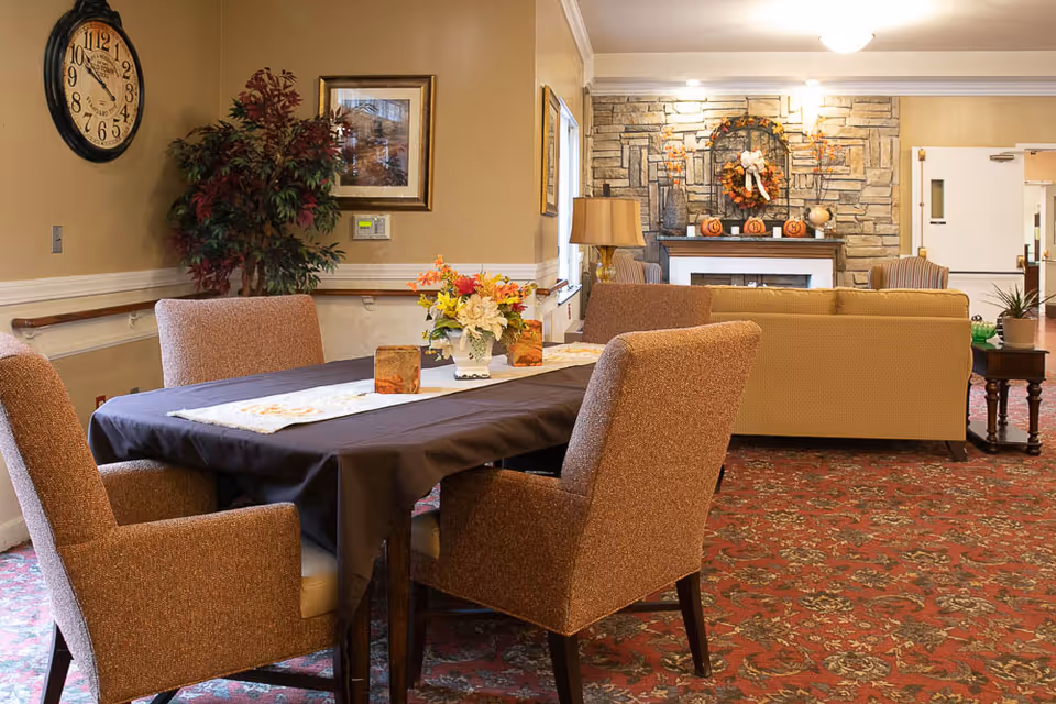 A cozy interior room with a table covered by a dark tablecloth and four upholstered chairs around it. A floral centerpiece and decorative items are on the table. In the background, there is a stone fireplace decorated with autumn-themed wreath and pumpkins, a beige sofa, and two armchairs. The room has warm lighting, a large wall clock, a framed picture, and a potted plant in the corner.