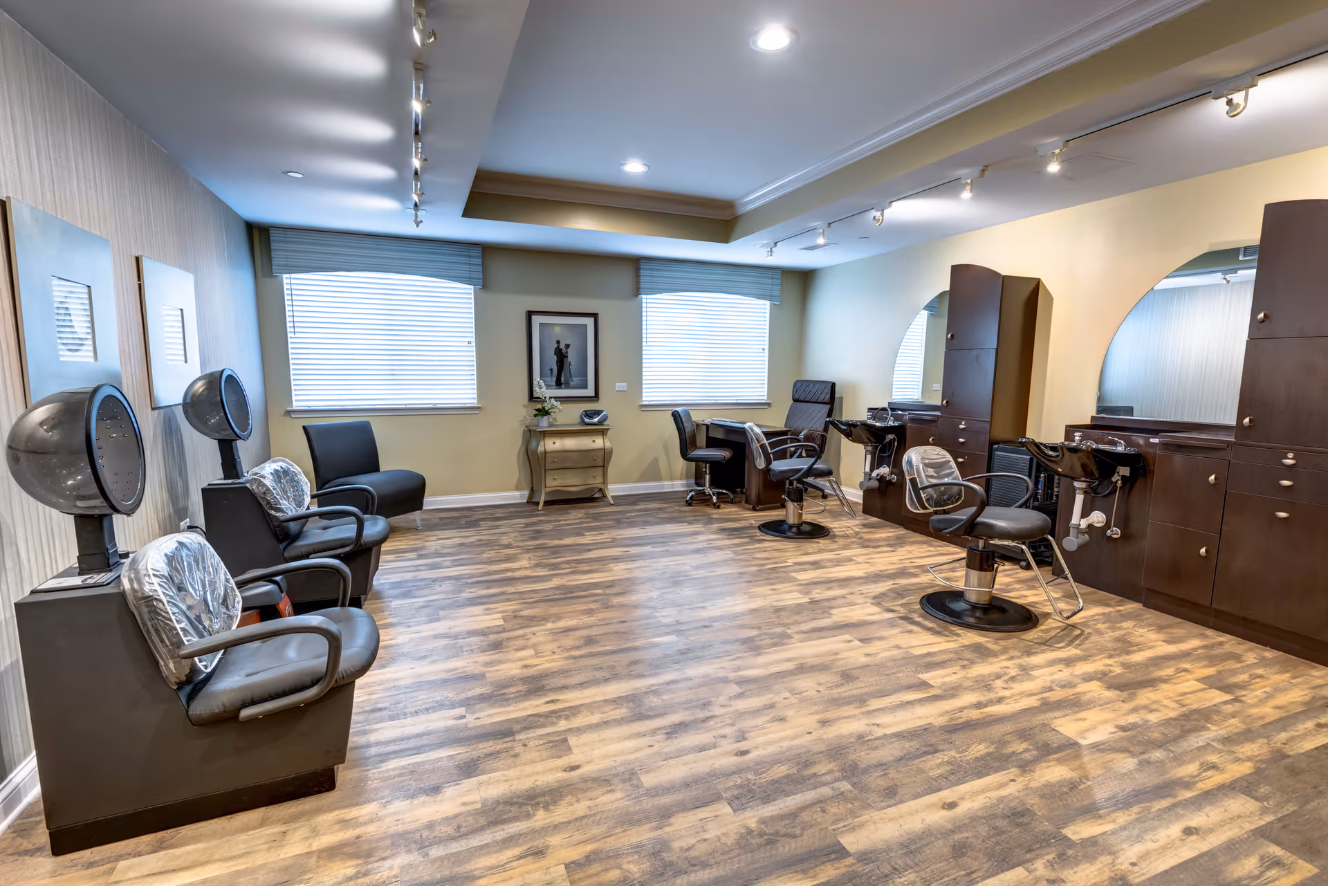 Interior view of a senior living facility's hair salon with wooden flooring, two hair drying chairs on the left, two styling chairs in front of mirrors on the right, and a small table with a framed picture and a flower vase between two windows with blinds.