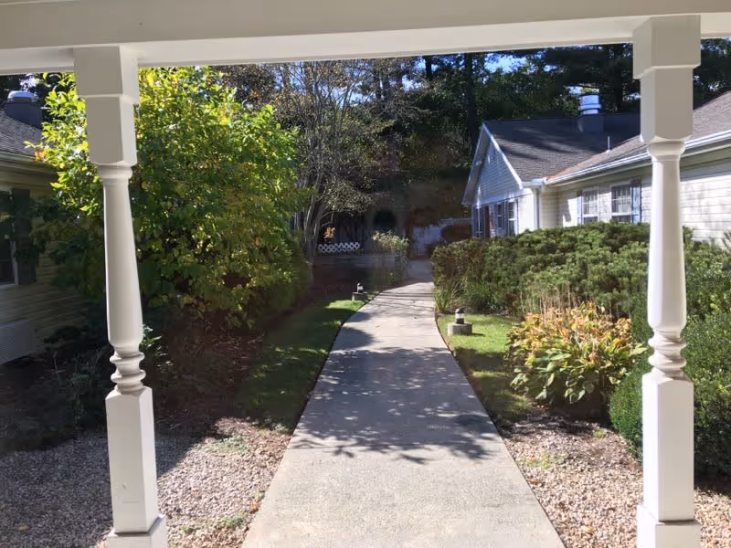 View of a paved walkway flanked by bushes and plants leading between two single-story buildings under a clear sky, framed by white porch columns.