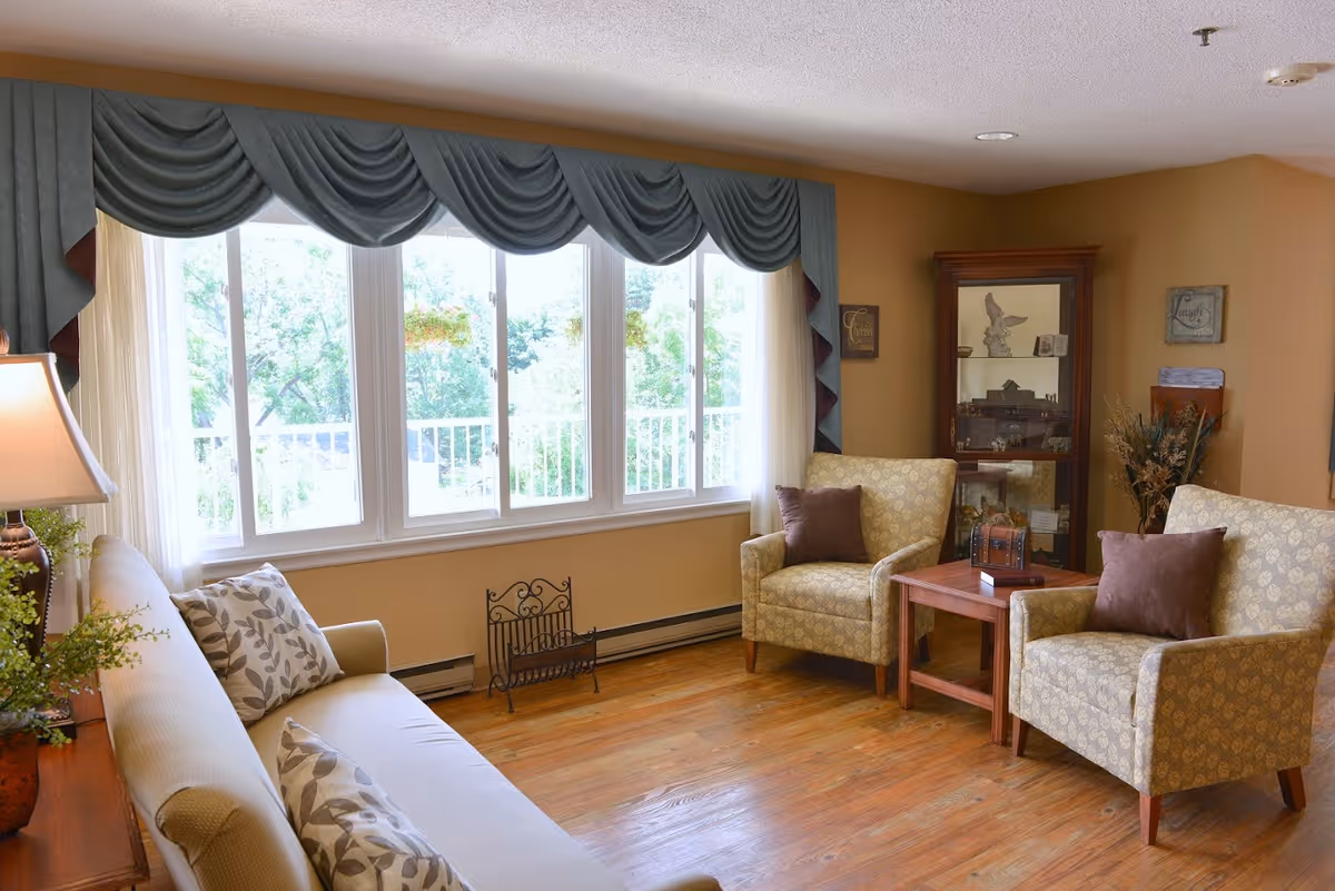 A cozy living room with a large window covered by blue valance curtains, a beige sofa with patterned pillows, two patterned armchairs with brown cushions, a wooden side table between the chairs, a wooden display cabinet in the corner, and a lamp on a wooden table next to the sofa. The room has wooden flooring and warm beige walls.