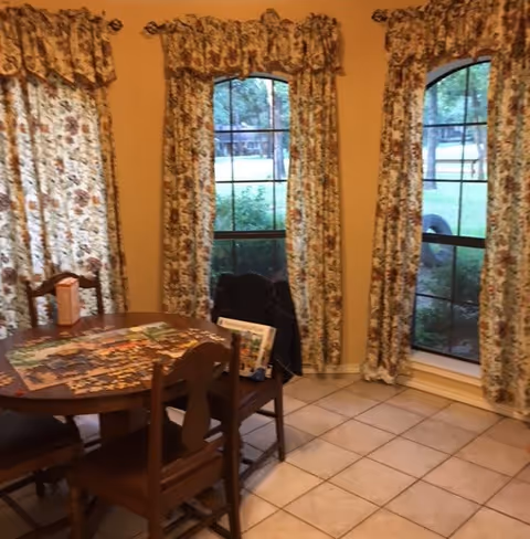 A cozy room with three tall windows covered by floral-patterned curtains. In front of the windows is a wooden table with four matching chairs. On the table, there is a partially completed jigsaw puzzle and a box, with a jacket draped over one chair. The floor is tiled with light-colored square tiles.