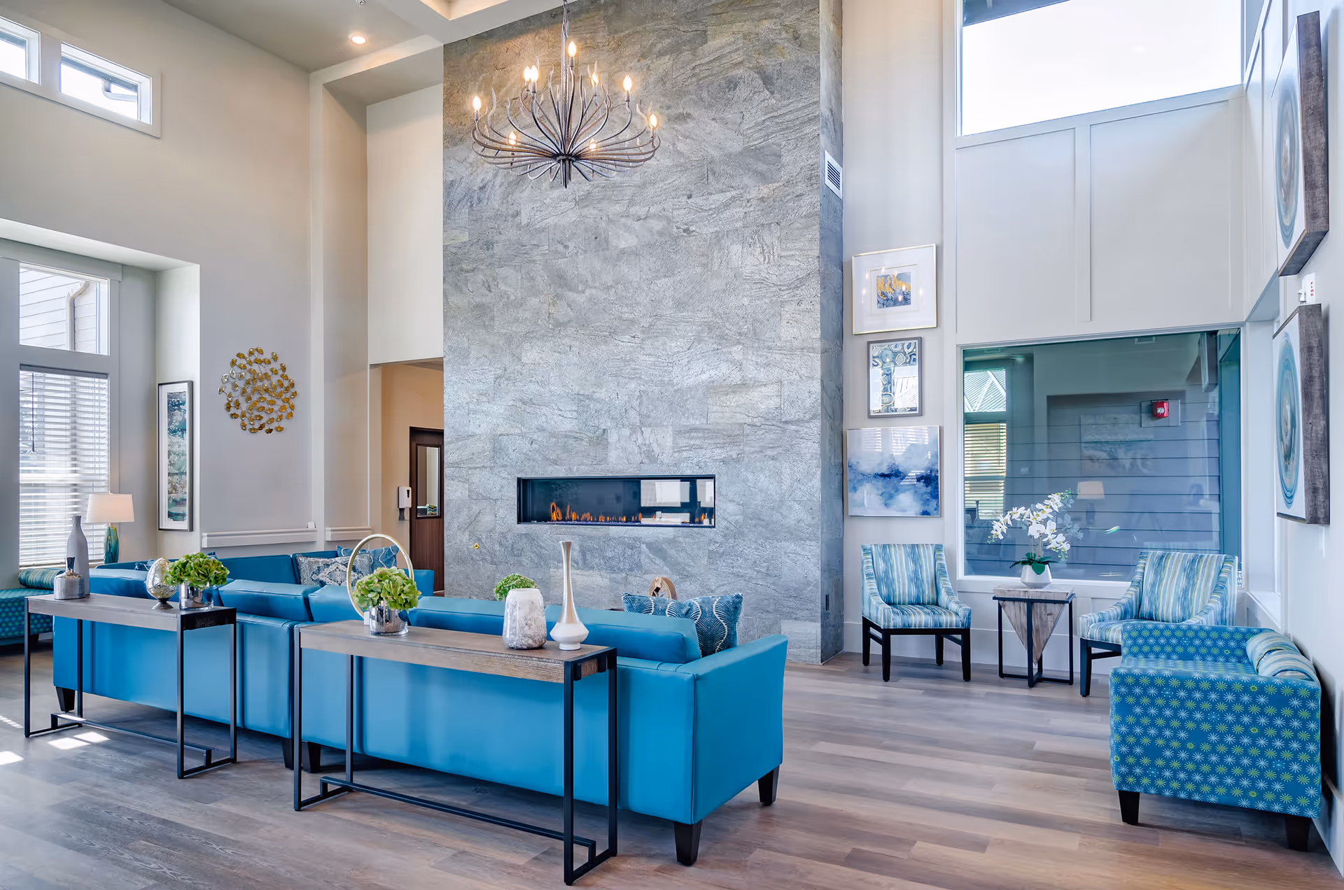 Bright modern communal living room with teal sofas, patterned chairs, a tall stone fireplace, and decorative lighting.