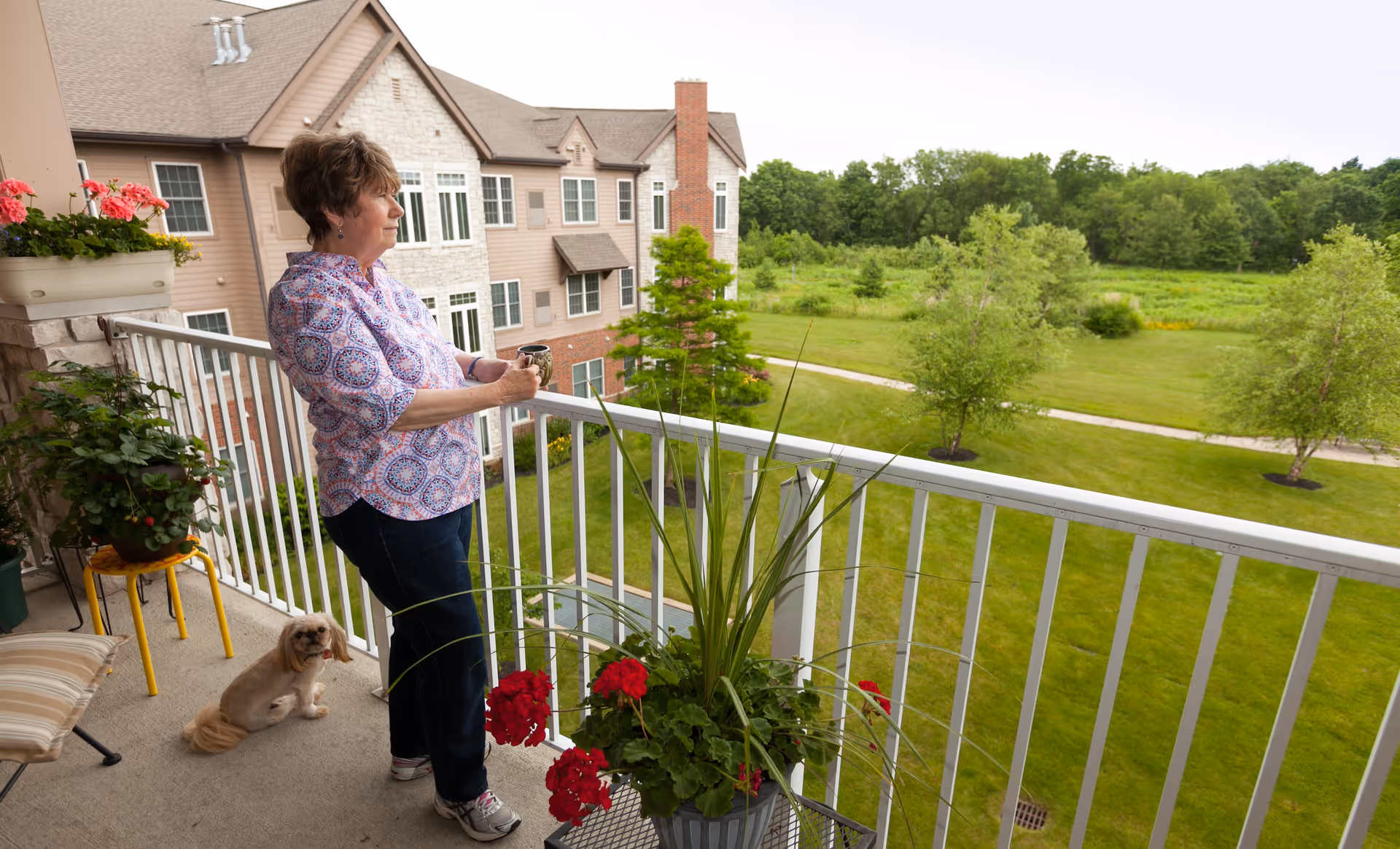 An older woman stands on a balcony holding a mug, looking out over a green lawn and trees. A small dog sits on the balcony floor near her. The balcony has potted plants with red flowers and a chair with a striped cushion. In the background, there is a multi-story building with beige and stone exterior walls.