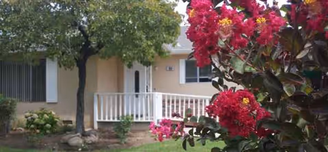 Single-story senior living facility exterior with a front porch, white railing, and pink flowers.