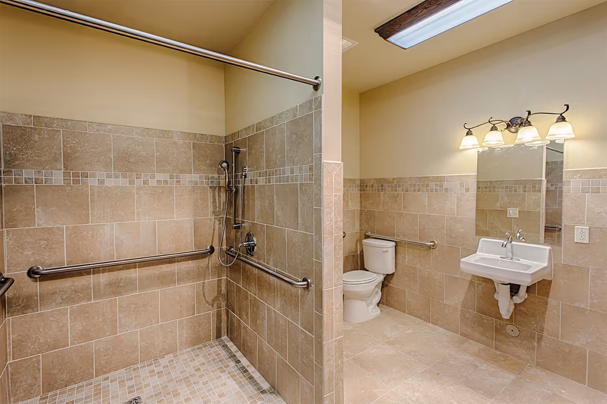 A spacious bathroom with beige tiled walls and floor. The bathroom features a walk-in shower with a handheld showerhead and multiple grab bars for accessibility. There is a white toilet and a wall-mounted sink with a mirror above it, illuminated by a four-light fixture.