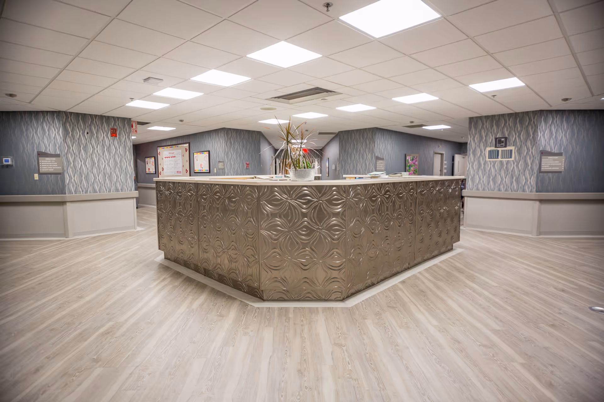A spacious reception or nurse station area in a senior living facility with a large, decorative metallic counter in the center. The floor is light wood, and the walls have a textured gray wallpaper. The ceiling has multiple recessed lights, and there are informational boards and signs on the walls.