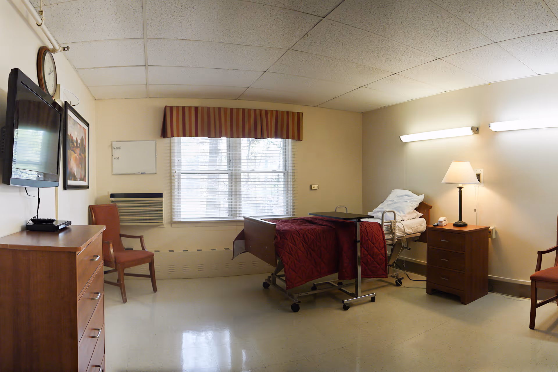 A simple, clean senior living facility bedroom with a hospital-style bed covered with a red quilt, a wooden nightstand with a lamp and phone, two chairs, a wall-mounted TV, a clock, and a window with blinds and a striped valance.