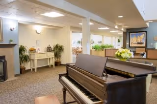Interior view of a senior living facility common area featuring a grand piano in the foreground, a fireplace on the left, plants, a small white console table with decorative items, and a dining area with tables and chairs in the background. The space is well-lit with ceiling lights and decorated with framed artwork and flowers.